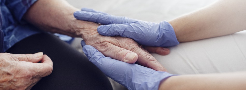 En omsorgsgivare håller i en äldre persons hand med båda sina händer iklädda gummihandskar.