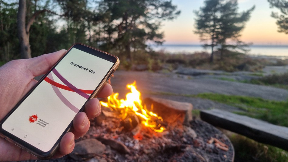 Hand håller en mobiltelefon med appen Brandrisk ute. I bakgrunden en grillplats vid en sjö.