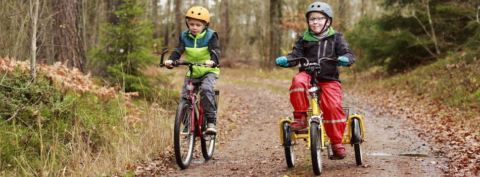 Tvillingpojkar som cyklar på skogsväg. Ena pojken har en funktionsvariation  och cyklar på trehjulig cykel.