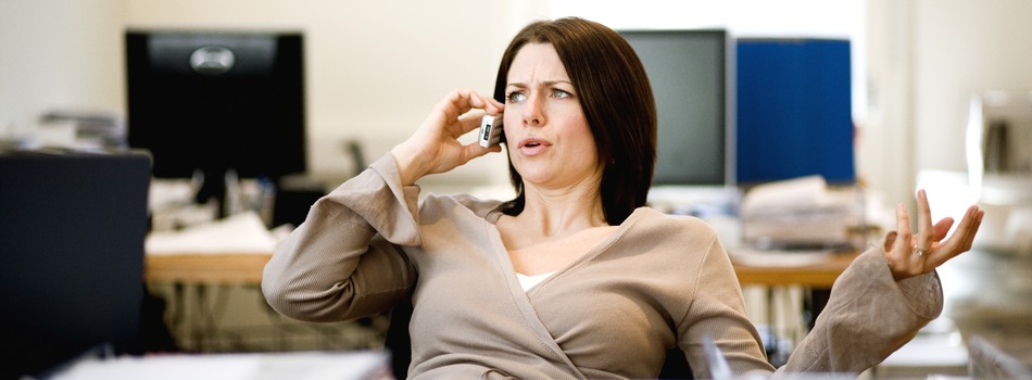 Mörkhårig kvinna sitter vid ett skrivbord och pratar i telefon med högerhanden och gestikulerar med sin vänstra hand